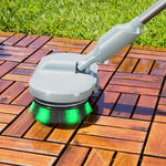 A close-up of a cleaning brush with a gray handle and green bristles, cleaning a brown wooden deck outdoors next to green grass.