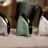Three compact hair styling tools with curved ergonomic designs are lined up on a textured gray surface. From left to right, the colors are metallic silver, matte sage green, and glossy white, each featuring a black accent panel and a small white button.
