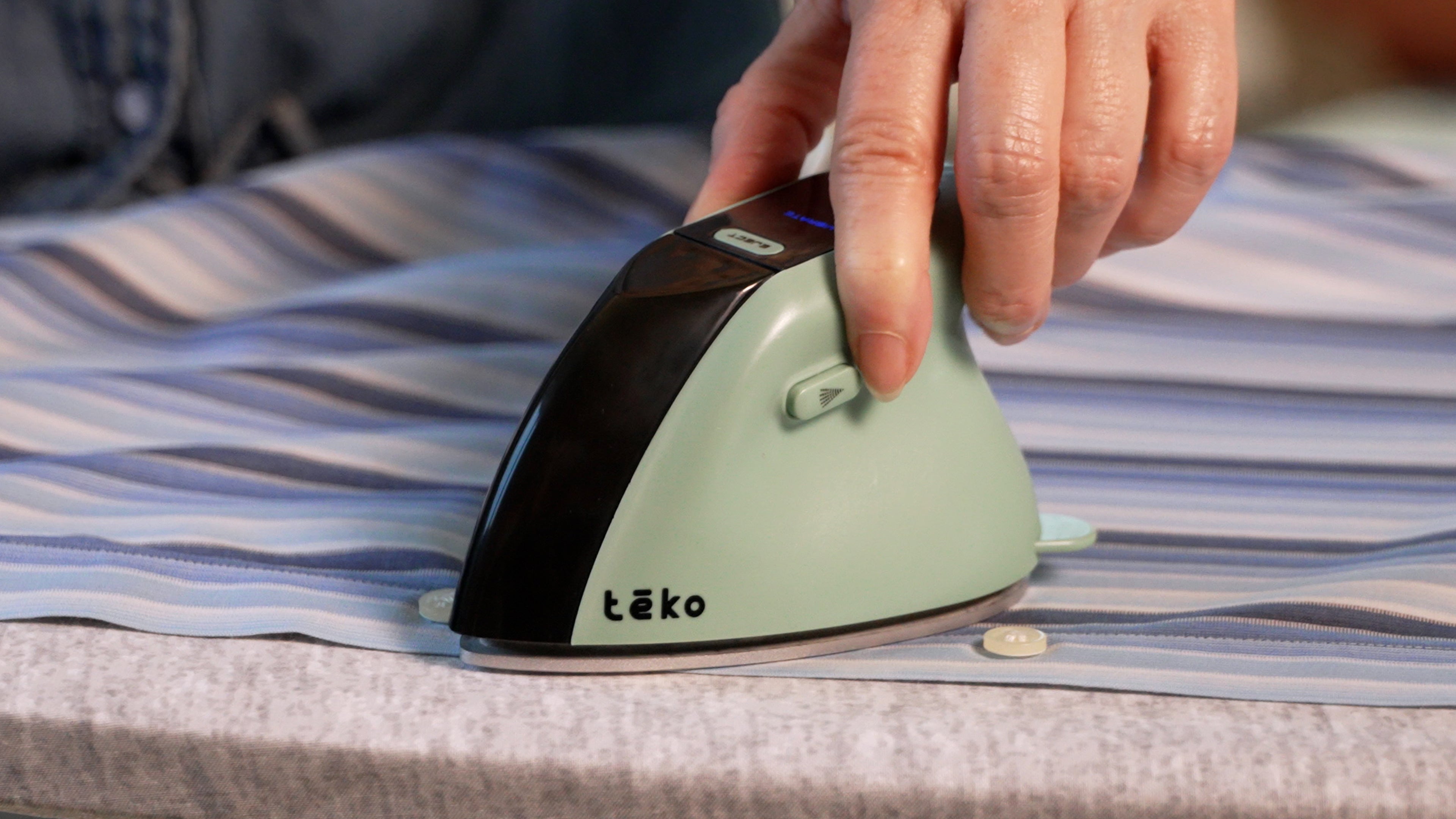 A close-up of a hand using a Teko portable steam iron on a striped blue and white shirt over a gray ironing board.