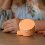 A small round peach-colored portable power bank or charging case sits on a wooden table, with a person in a floral blouse blurred in the background.