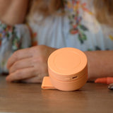 A small round peach-colored portable power bank or charging case sits on a wooden table, with a person in a floral blouse blurred in the background.