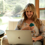 A woman sitting in a beige armchair in front of large windows, holding a small teal circular object and looking at a silver laptop. She is wearing a light floral blouse.