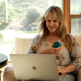 A woman sitting in a beige armchair in front of large windows, holding a small teal circular object and looking at a silver laptop. She is wearing a light floral blouse.