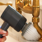 A hand holding a gray and black palm scrubber with a brush attachment, turning on a faucet to demonstrate its use.