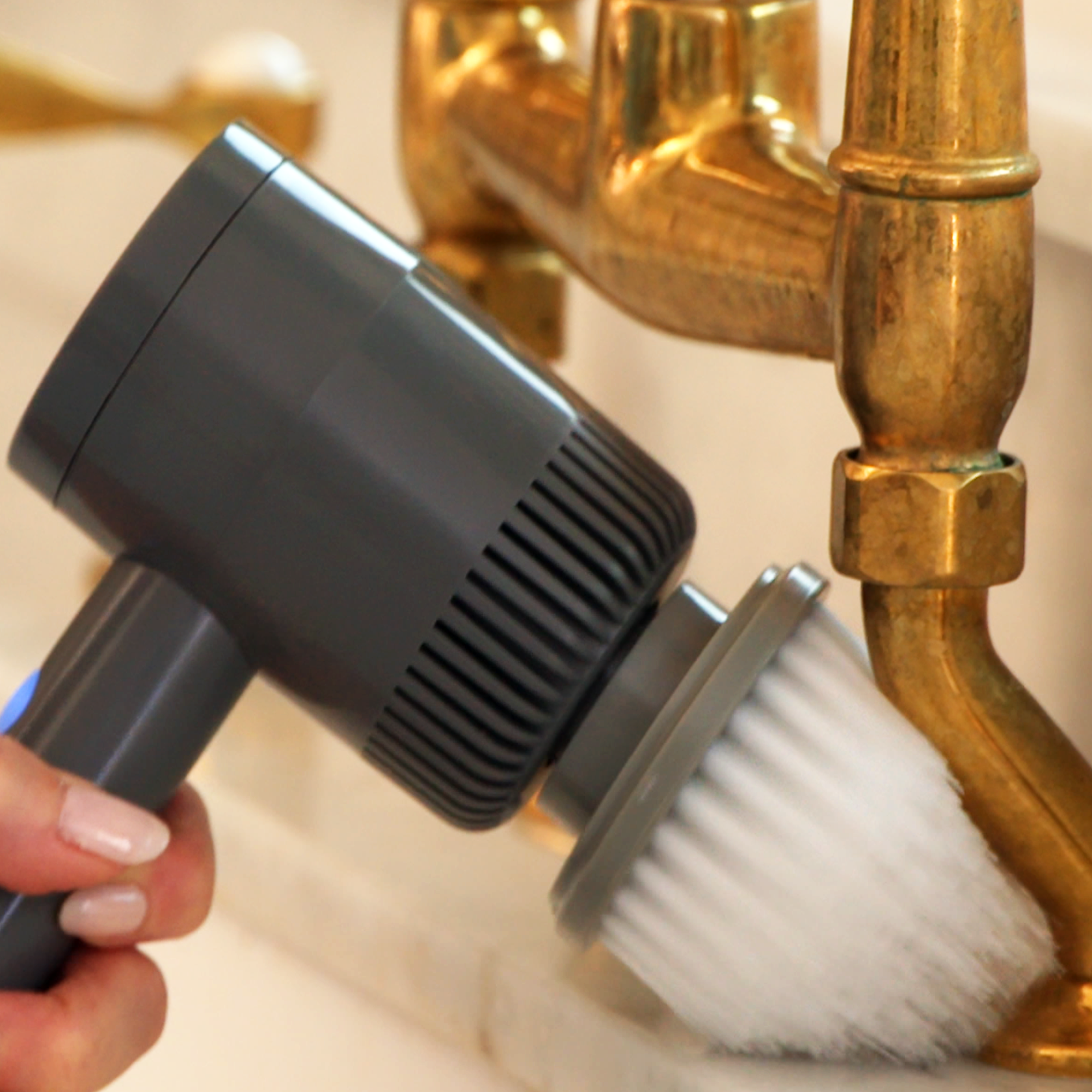 A hand holding a gray and black palm scrubber with a brush attachment, turning on a faucet to demonstrate its use.